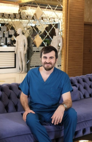 A person with a beard and wearing a blue V-neck shirt smiles in front of a decorative background featuring reflective surfaces that could easily belong in a surgeons waiting room, complete with abstract sculptures hinting at the artistry behind each necklift.